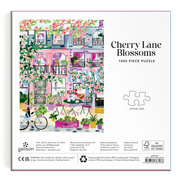Sabina Fenn’s Cherry Lane Blossoms 1000 Piece Puzzle features a floral scene with a pink building, balconies, trees, potted plants, a café table and chairs, and a white bicycle.