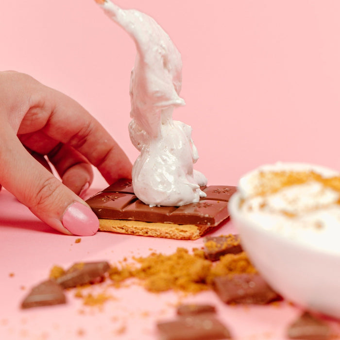 A hand holding the base of a s'more which is a graham cracker and bar of chocolate while it scoops gimmie s'more marshmallow on top of the chocolate with a spoon