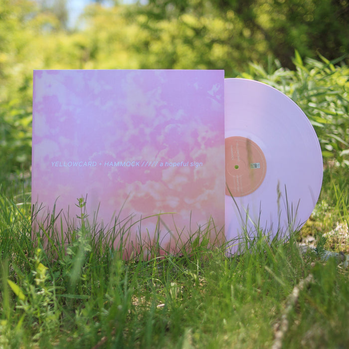 A pastel pink Yellowcard LP vinyl and matching album cover with soft pink clouds and a hopeful sign text rest on green grass outdoors, surrounded by sunlight and foliage.