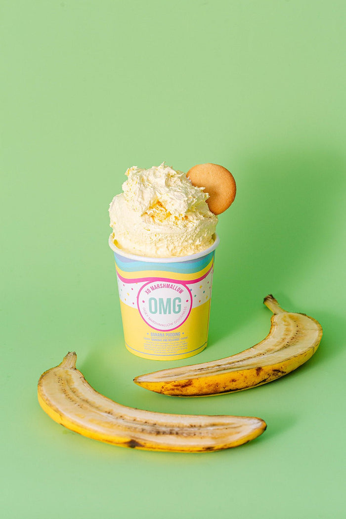 A pint of Banana pudding flavored XO Marshmallow ooey marshmallow goodness with a nilla wafer sticking out of the top of it against a light green background. There is a banana cut in half laying in front of it.
