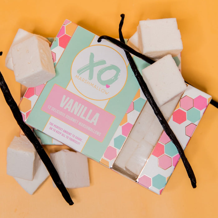 A box of vanilla gourmet marshmallows against an orange background with vanilla beans and vanilla marshmallows around it