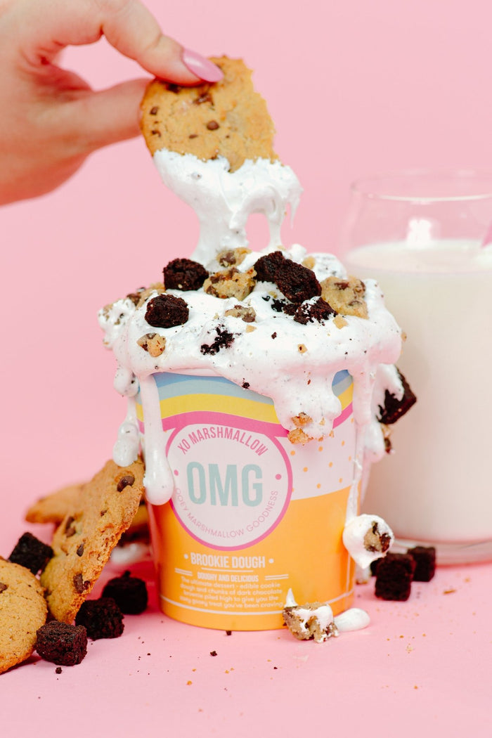 A person dipping a chocolate chip cookie into a pint of XO Marshmallow Brookie Dough ooey marshmallow goodness with chunks of brownie and cookie dough overflowing the pint. There is a glass of milk in the background.