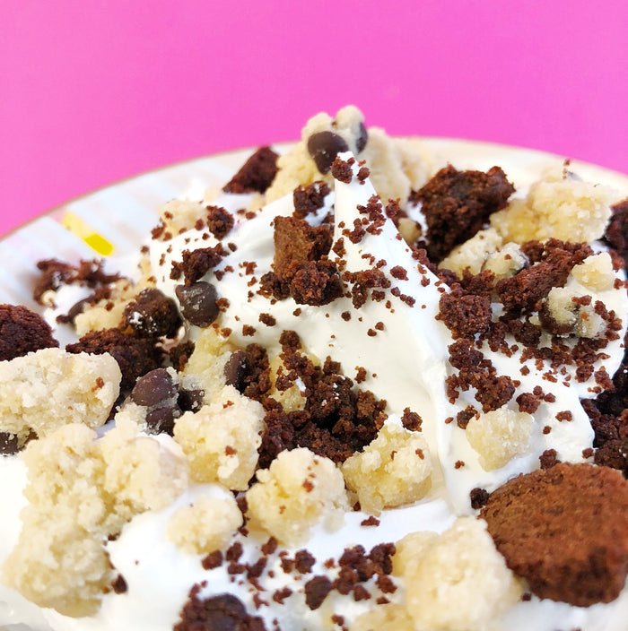 A photo of the gourmet marshmallow brookie dough fluff inside of a bowl against a pink background. There are lots of edible cookie dough and brownie pieces on top.