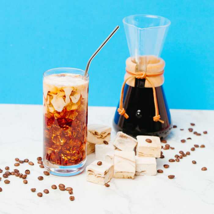 A cup of iced coffee, a carafe of coffee and caffeinated marshmallows against a blue background