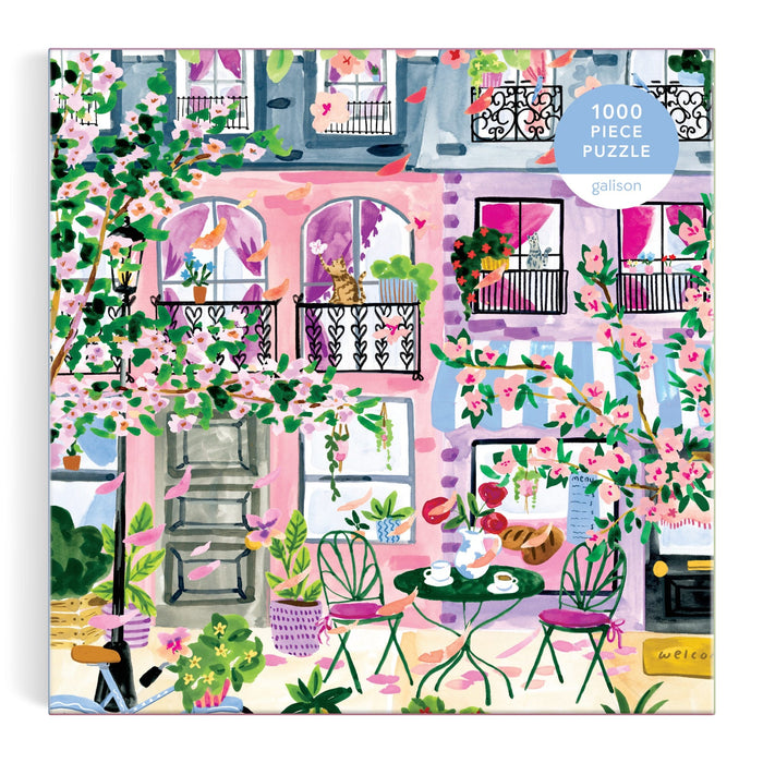 Cherry Lane Blossoms 1000 Piece Puzzle by Sabina Fenn features a vibrant pink building with balconies, blooming flowers, lush greenery, and a café table with green chairs on a patio—perfect for springtime street scene lovers.