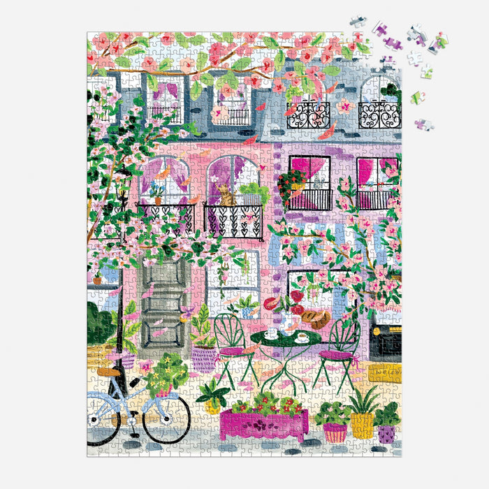 The Sabina Fenn Cherry Lane Blossoms 1000 Piece Puzzle features a vibrant building with pink walls, flowering trees, potted plants, a bicycle, and a café table on the sidewalk. A few pieces remain unfinished in the top right corner.
