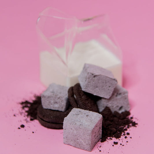 A clear plastic milk carton full of milk next to Cookies and Cream marshmallows, with loose chocolate sandwich cookie pieces and cocoa powder scattered around, presented on a pink background.