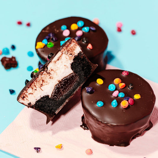 Two brownie cups topped with chocolate ganache and multicolored rainbow chips on a blue background with additional chips scattered around.