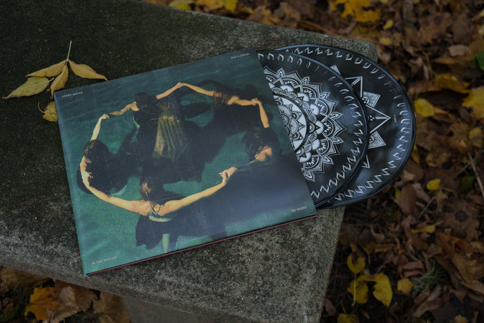 A Limited Edition Vinyl by Night Verses with a black and white geometric design partially slides out of its album cover, which shows women in black dresses holding hands in a circle, all resting on a stone bench amid fallen autumn leaves.