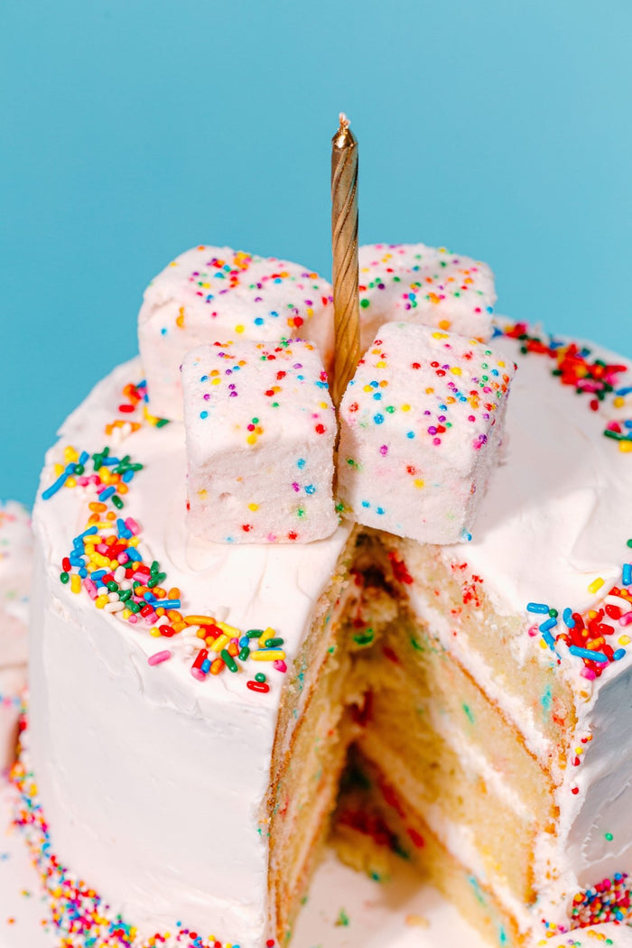 A white birthday cake covered in sprinkles with a giant slice of cake missing with XO Marshmallow Funfetti homemade marshmallows on top with one gold birthday candle in the middle. It shows that the Funfetti flavor is birthday cake.