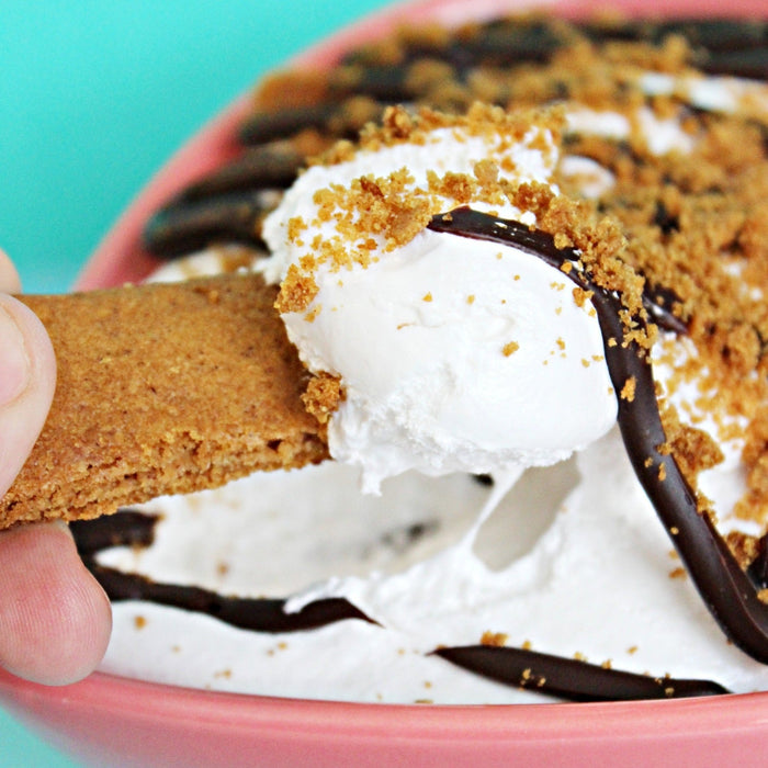 A bowl of gimmie s'more gourmet marshmallow spreadable creme topped with chocolate ganache and crushed gluten free graham crackers. A hand is using another graham cracker to dip it into the fluff