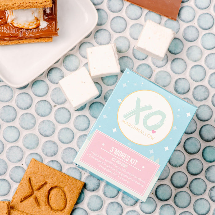 A gourmet s'mores kit with marshmallows, graham crackers, and chocolate bars laid out on a blue patterned surface, with a box labeled 'XO' visible.
