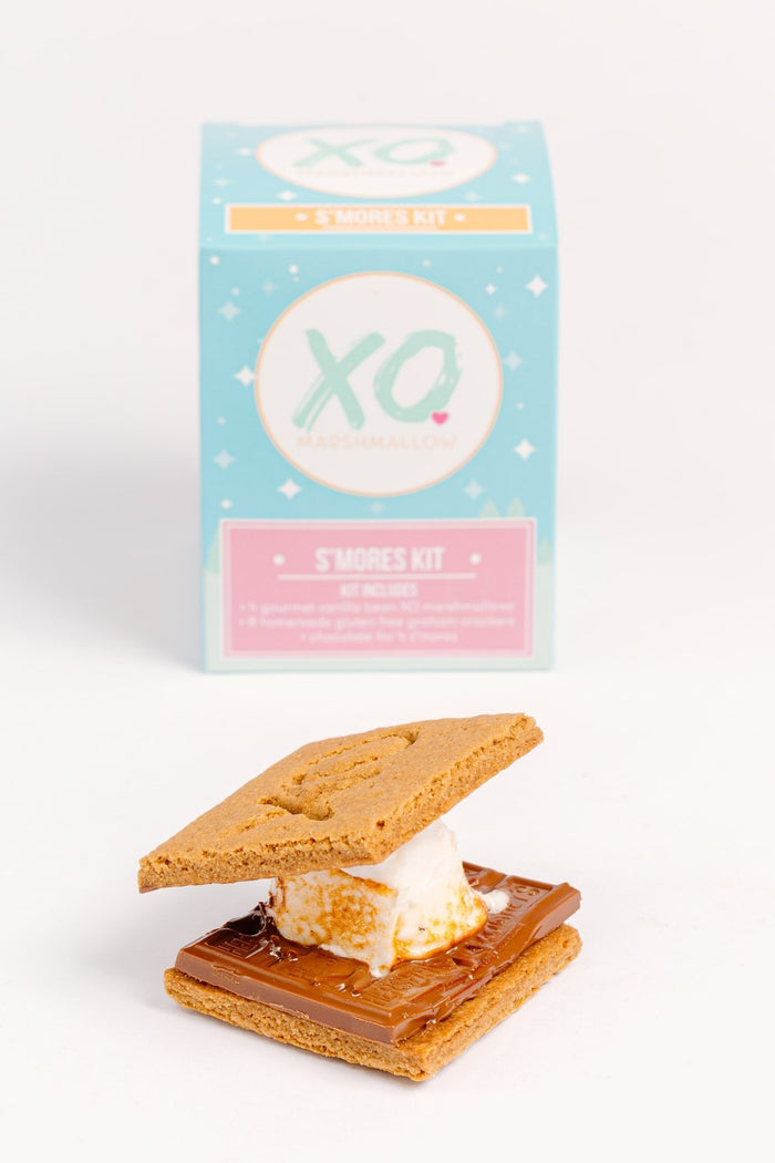 A gourmet xo marshmallow s'more in front of the s'mores kit box in comes in. The box shows that it's perfect for gifting, corporate gifting, client gifting and more
