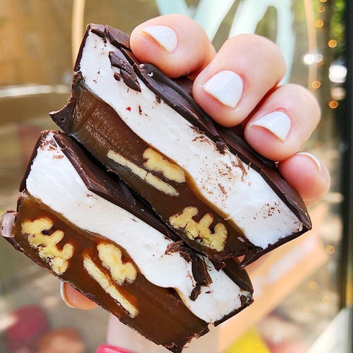 A hand holding a cross section of XO Marshmallow gourmet turtles with layers of caramel with pecans mixed in and topped with gourmet vanilla marshmallow and everything is dipped in chocolate