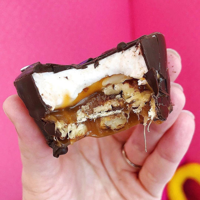 A hand holding a marshmallow turtle candy that has had a bite taken out of it. The caramel sauce is dripping down out of it onto the hand