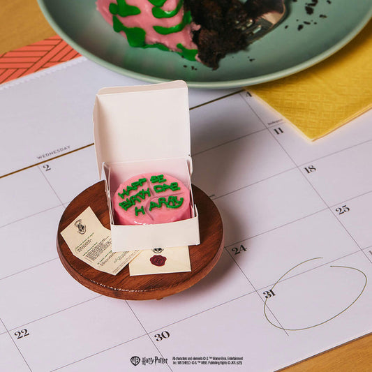 Pink cake with green text on a wooden plate, calendar in the background