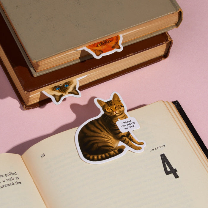 The Brass Monkey Moody Cats Magnetic Bookmark Set features three cat bookmarks peeking from books—one with a sticker saying, "I heard the villain sucked." A fun, light pink set perfect for cat-loving readers.