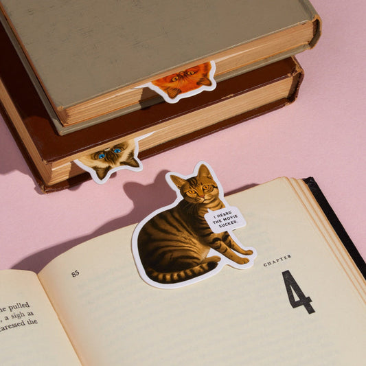 The Brass Monkey Moody Cats Magnetic Bookmark Set features three cat bookmarks peeking from books—one with a sticker saying, "I heard the villain sucked." A fun, light pink set perfect for cat-loving readers.
