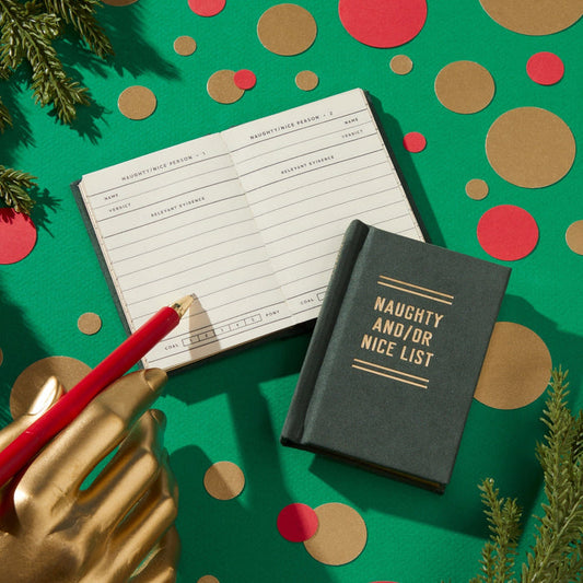 A gold mannequin hand holds a red pencil over an open Naughty and/or Nice Tiny Diary by Brass Monkey, set on a green background with festive accents. A closed pocket-sized version of the diary lies nearby, both featuring the playful title.