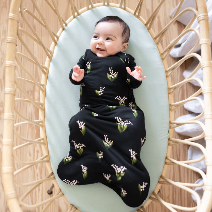 Infant laying on Kyte Baby Oval Bassinet Sheet in Sage wearing the 1.0 Sleep Bag in Midnight Lily