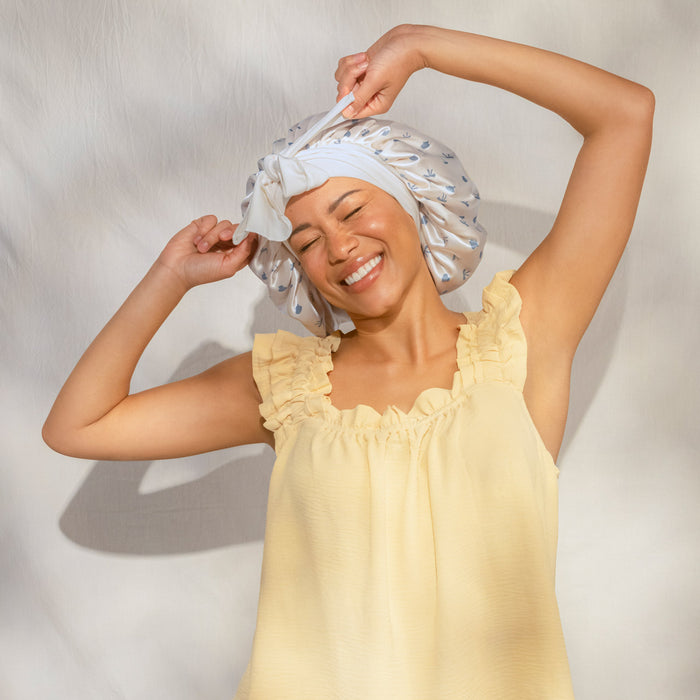 Smiling female model in yellow ruffled top wearing KITSCH Pearlized Cream & Coastal Blue Print Oversized Satin Bonnet — arms raised, joyful lifestyle portrait shot — Adjustable, double-layered satin — For preserving your hair overnight