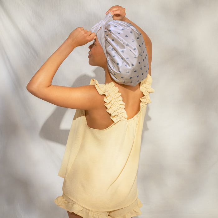 Female model in yellow ruffled top, back/side view, adjusting KITSCH Pearlized Cream & Coastal Blue Print Oversized Satin Bonnet on head — hands on bonnet, lifestyle portrait shot — Adjustable, double-layered satin with seashells & bows print — For preserving your hair overnight
