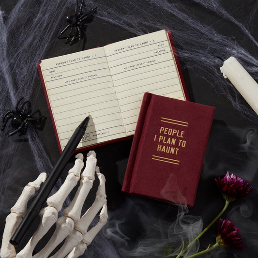 A spooky Halloween scene features the Brass Monkey People I Plan To Haunt Tiny Diary, an open page with a pen in a skeleton hand, fake spiders, cobwebs, a white candle, and dark purple flowers on a black surface.