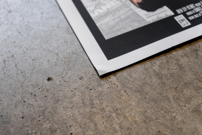 A close-up of a slightly damaged Bane 18x24 poster with a black border on gray concrete. The bottom corner is lifted, revealing text and a QR code, with no refunds faintly visible.