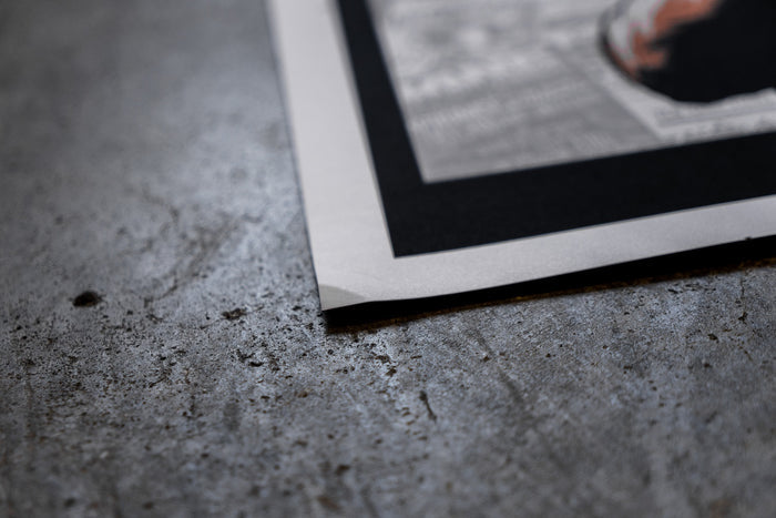 A close-up of a slightly damaged Bane 18x24 poster on textured gray concrete. The edge is slightly bent, and the image features a white and black border.