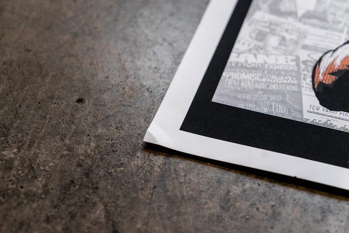 A close-up of the corner of a slightly damaged Bane 18x24 poster on a textured gray surface, featuring part of a black-and-white design with some text and color; no refunds.