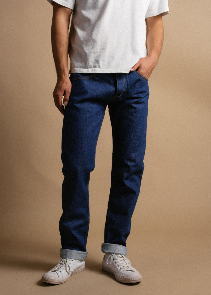 Man in white shirt, blue jeans, and white sneakers stands casually against a beige background, hands in pockets.