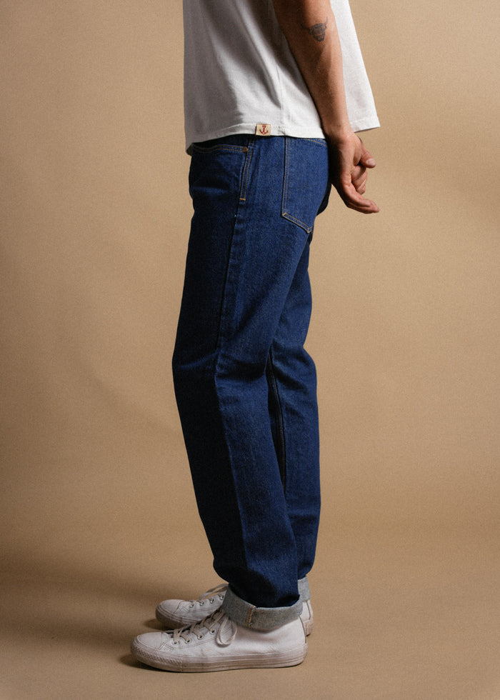 Man in blue jeans and white sneakers stands on a beige background, wearing a white shirt with rolled-up sleeves.