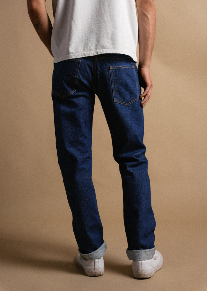 Person in blue jeans, white sneakers, and a white shirt stands against a beige background, facing away, hands relaxed by sides.