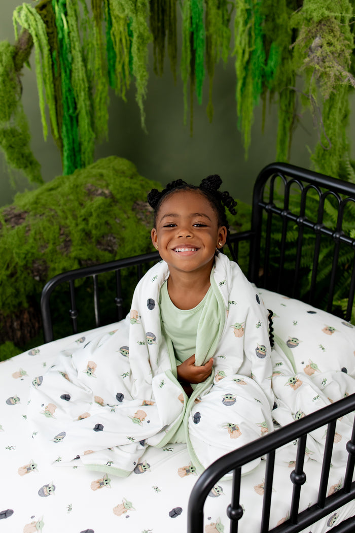 Smiling girl sitting in bed with the Toddler Blanket in Grogu™ 1.0 around her shoulders