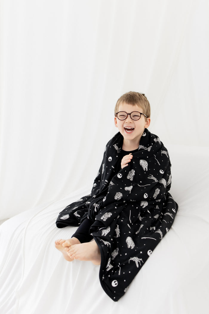 Young smiling boy sitting on a white blanket with the Toddler Blanket in STAR WARS™ 1.0 around his shoulders