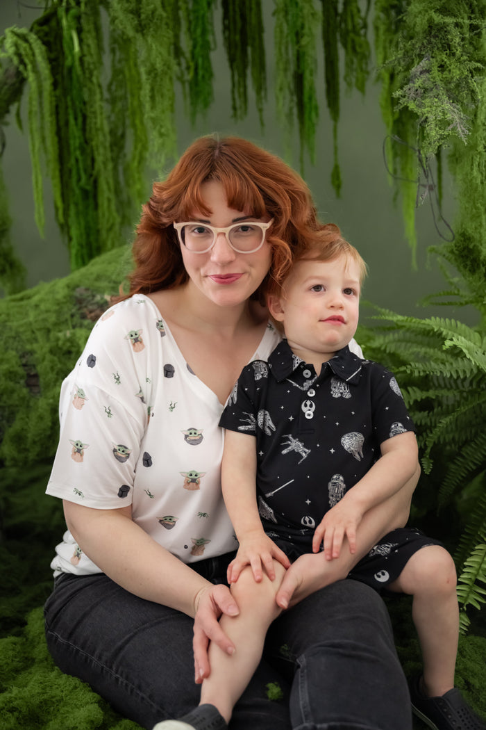 Young boy wearing the Toddler Short Sleeve Polo in STAR WARS™ sitting on his mother's lap who is wearing the Women's V-Neck in Grogu™