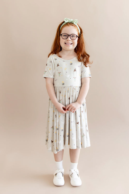 Child wearing a Harry Potter patterned twirl dress with a green headband and white socks and shoes on a beige background
