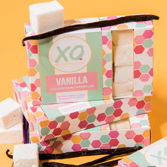 A close up photo of 3 Boxes of XO Marshmallow vanilla marshmallows stacked up in their packaging. Vanilla beans and vanilla marshmallows are scattered about the photo. Everything is against an orange background.
