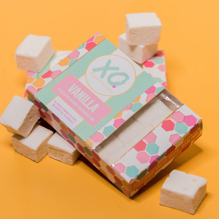 One box of XO Marshmallow Gourmet Vanilla Marshmallows laying flat against an orange background. Vanilla marshmallows are scattered around it.