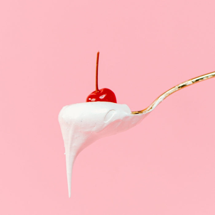 A gold spoon with a scoop of XO Marshmallow Vanilla Marshmallow fluff dripping off of it. There is a cherry on top of it. The cherry is only for the photo and not part of the product. It's all against a light pink background.