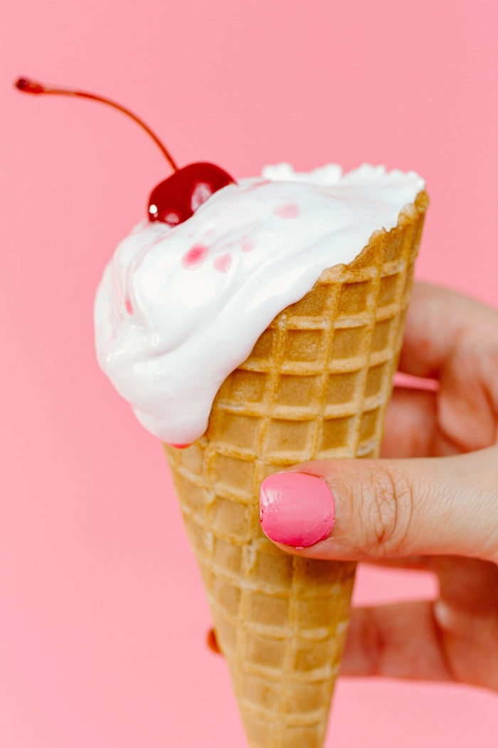 A hand with pink nail polish holds a waffle ice cream cone filled with XO Marshmallow vanilla marshmallow creme topped with a cherry against a light pink background.
