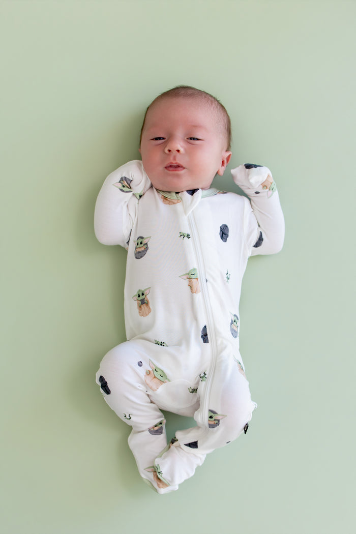 Newborn laying on a light green surface wearing the Zippered Footie in Grogu™ with the fold over hand cuffs over the baby's hands